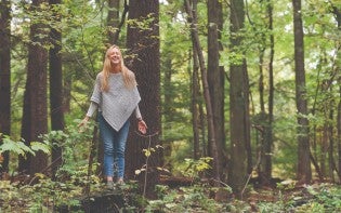 Smiling woman with arms lowered and eyes closed standing in the woods.