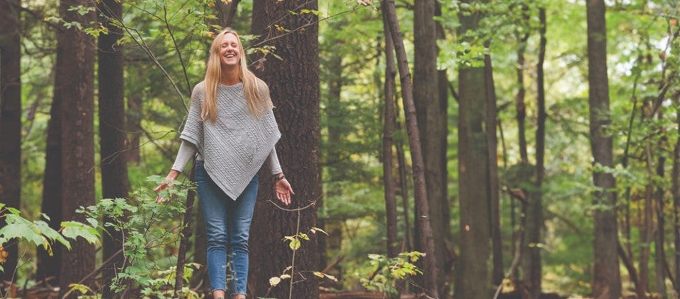 Smiling woman with arms lowered and eyes closed standing in the woods.